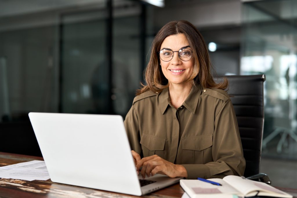 Feliz e sorridente mulher de negócios madura e de meia-idade, investidora, gerente, executiva ou advogada, consultora, olhando para a câmera no local de trabalho, trabalhando em um computador portátil no escritório, retrato.