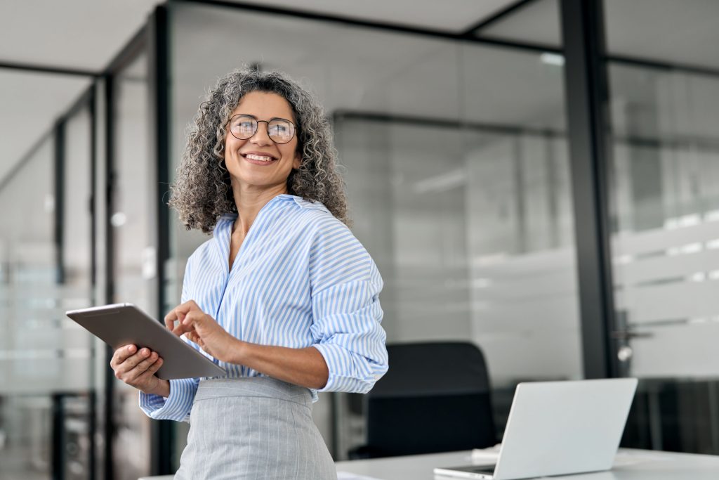 Sorridente profissional de negócios madura, gerente de banco, executiva feliz ou empresária segura segurando um tablet digital, em pé no escritório, olhando para longe para um espaço em branco.