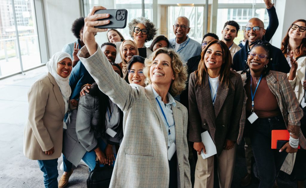 Um grupo diverso de profissionais tirando uma selfie alegre juntos durante um seminário ou workshop. Eles estão sorrindo e parecem engajados.